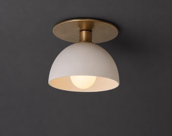 Pode incluir: Uma luminária de teto de montagem embutida com uma base circular de latão e uma cúpula branca fosca. A luminária inclui uma lâmpada central que emite um brilho quente, iluminando o interior da cúpula branca.