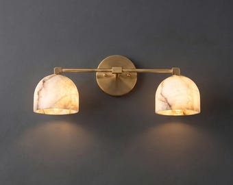 May include: A two-light wall sconce featuring a horizontal brass bar and a circular wall mount. The fixture includes two dome-shaped shades made of translucent, marbled stone with warm, veined patterns that emit a soft, diffused light.