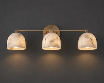 May include: A three-light wall fixture featuring a horizontal brass bar and a circular wall mount. Three dome-shaped shades made of white and grey marbled stone are attached to the bar, each housing a single light bulb that emits a warm glow.