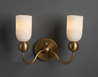 Pode incluir: Um candeeiro de parede de duas luzes com uma placa circular de latão antigo e dois braços curvos que se estendem para fora. Cada braço suporta um detalhe esférico de latão e um abajur cilíndrico de pedra branca translúcida.