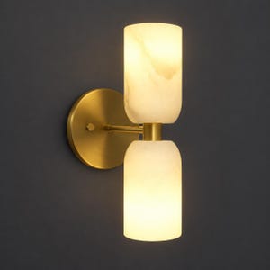 May include: A wall sconce featuring a circular brushed brass backplate and a horizontal arm supporting two vertically stacked, cylindrical, translucent white stone shades that emit a warm, soft glow from the internal light source.
