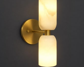 Pode incluir: Um candeeiro de parede com uma placa circular de latão escovado e um braço horizontal que suporta dois abajures cilíndricos de pedra branca translúcida empilhados verticalmente, que emitem um brilho quente e suave da fonte de luz.