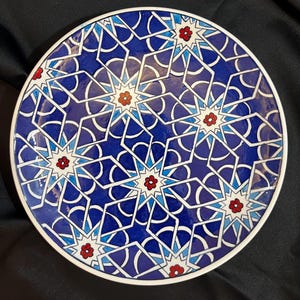 Może przedstawiać: Okrągły ceramiczny talerz z misternym geometrycznym wzorem splecionych białych linii na głębokim niebieskim tle. Projekt zawiera powtarzające się kształty gwiazd z małymi czerwonymi i pomarańczowymi kwiatowymi środkami oraz białą krawędzią.