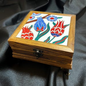 May include: A square wooden box with a hinged lid featuring a ceramic tile inlay. The tile displays a colorful floral pattern with red and blue tulips, green stems, and small blue flowers. The box has a dark metal latch and small decorative feet.