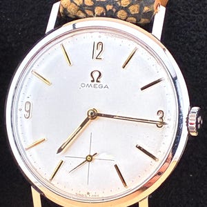 May include: A vintage Omega wristwatch with a silver face, gold hands, and gold hour markers. The watch has a leather band with a patterned design. The watch face displays the Omega logo and the words "Swiss Made".