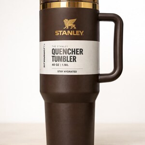 May include: A brown Stanley Quencher Tumbler with a handle. The tumbler has a gold rim and lid, and the Stanley logo is visible. The label reads "Quencher Tumbler" and "40 OZ | 1.18L". The text "Stay Hydrated" is also printed on the label.