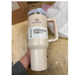 Puede incluir: Un vaso Stanley Quencher H2.0 de color beige claro con asa y tapa azul claro. El vaso tiene el logotipo de Stanley y las palabras "QUENCHER H2.0 TUMBLER" impresas. Es un vaso de 1,18L.