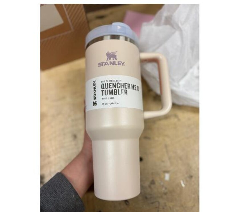 Puede incluir: Un vaso Stanley Quencher H2.0 de color beige claro con asa y tapa azul claro. El vaso tiene el logotipo de Stanley y las palabras "QUENCHER H2.0 TUMBLER" impresas. Es un vaso de 1,18L.