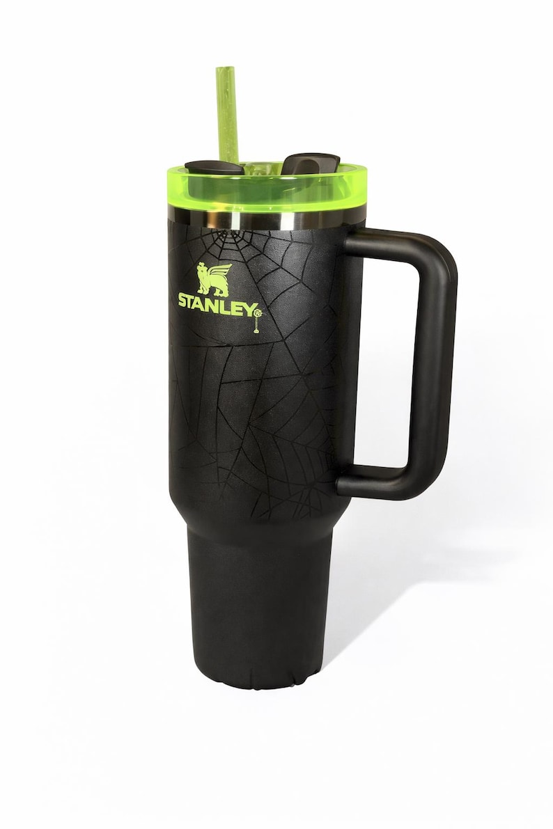 May include: Black Stanley tumbler with a neon green lid and straw. The tumbler has a handle and a spiderweb design. The Stanley logo is visible on the side.