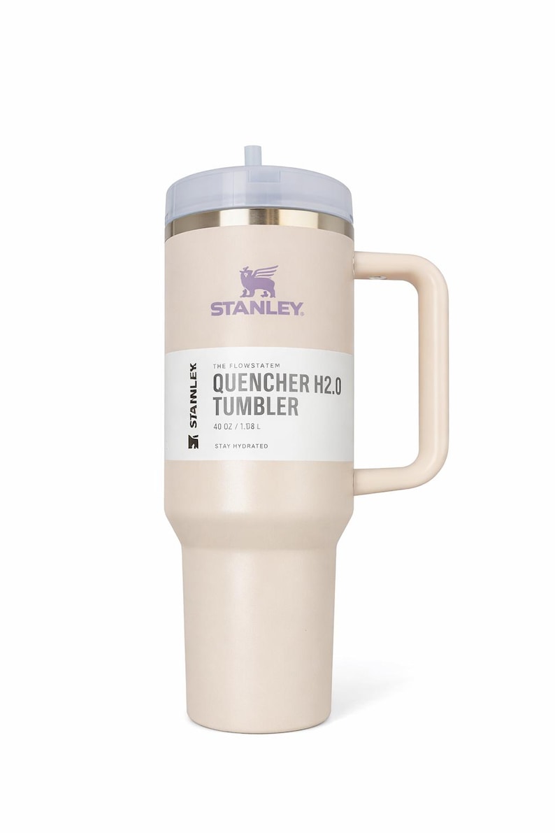Puede incluir: Vaso Stanley Quencher H2.0 de color crema con asa y tapa transparente. El vaso tiene el logotipo de Stanley y el texto "QUENCHER H2.0 TUMBLER" impreso. Contiene 1,18L. El texto "STAY HYDRATED" tambi&eacute;n est&aacute; impreso en el vaso.