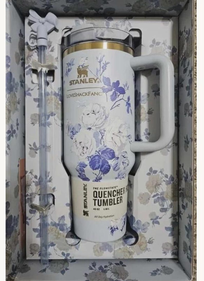 May include: A white Stanley Quencher H2.0 Flowstate tumbler with a 40 oz capacity, featuring a blue floral pattern, a gold rim, and a matching handle. The tumbler includes a clear straw with a white bow topper, all packaged in a floral box.