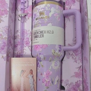 May include: A lavender Stanley Quencher H2.0 tumbler with a floral pattern, featuring a matching handle, lid, and a bow-adorned straw. The tumbler is housed in a floral-printed box alongside a card labeled LoveShackFancy, showing 40 oz / 1.18 L capacity.