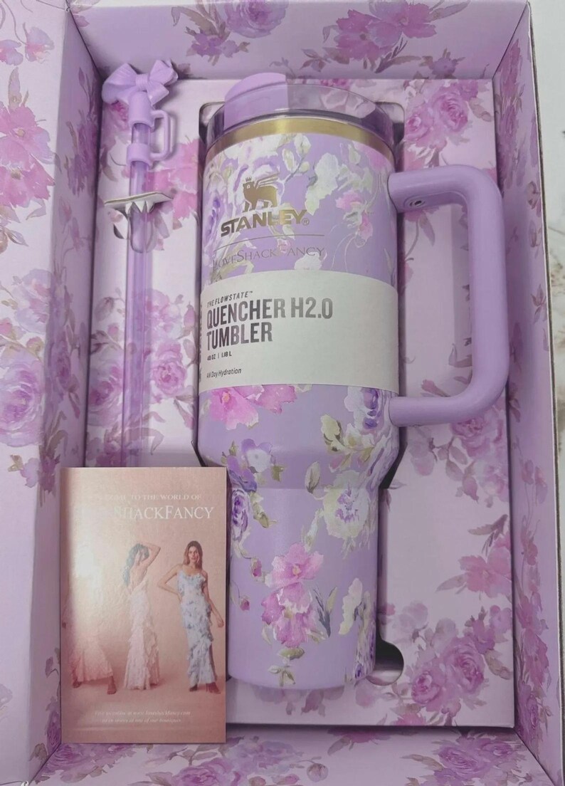 May include: A lavender Stanley Quencher H2.0 tumbler with a floral pattern, featuring a matching handle, lid, and a bow-adorned straw. The tumbler is housed in a floral-printed box alongside a card labeled LoveShackFancy, showing 40 oz / 1.18 L capacity.