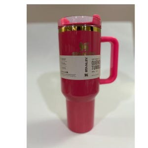 K&ouml;nnte beinhalten: Ein leuchtend pinkfarbener Stanley Quencher-Becher mit passendem Griff und Deckel. Der Becher hat ein goldenes Band am oberen Rand und ein wei&szlig;es Etikett mit schwarzem Text. Er fasst etwa 1,18 Liter.