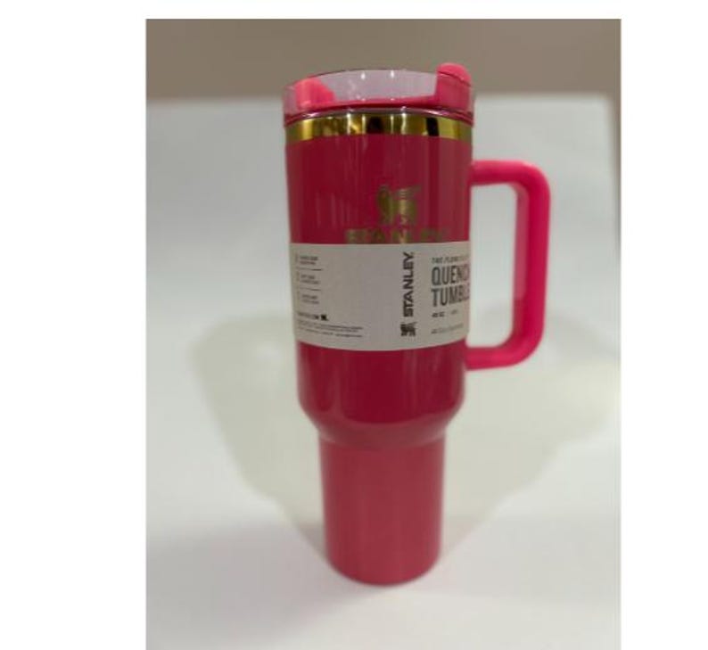 K&ouml;nnte beinhalten: Ein leuchtend pinkfarbener Stanley Quencher-Becher mit passendem Griff und Deckel. Der Becher hat ein goldenes Band am oberen Rand und ein wei&szlig;es Etikett mit schwarzem Text. Er fasst etwa 1,18 Liter.