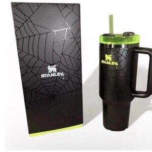 May include: Black Stanley tumbler with a handle and a green lid, next to a matching box. Both the tumbler and box feature a spiderweb design. The Stanley logo is visible on both items.