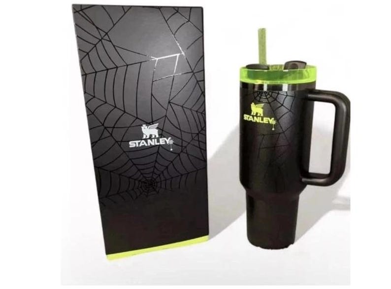 May include: Black Stanley tumbler with a handle and a green lid, next to a matching box. Both the tumbler and box feature a spiderweb design. The Stanley logo is visible on both items.