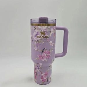 May include: A lavender stainless steel tumbler featuring a floral pattern with pink and purple roses. The cup has a gold-tone rim, a matching lavender handle, and a clear lid with a purple stopper. The Stanley and LoveShackFancy logos are printed in gold.