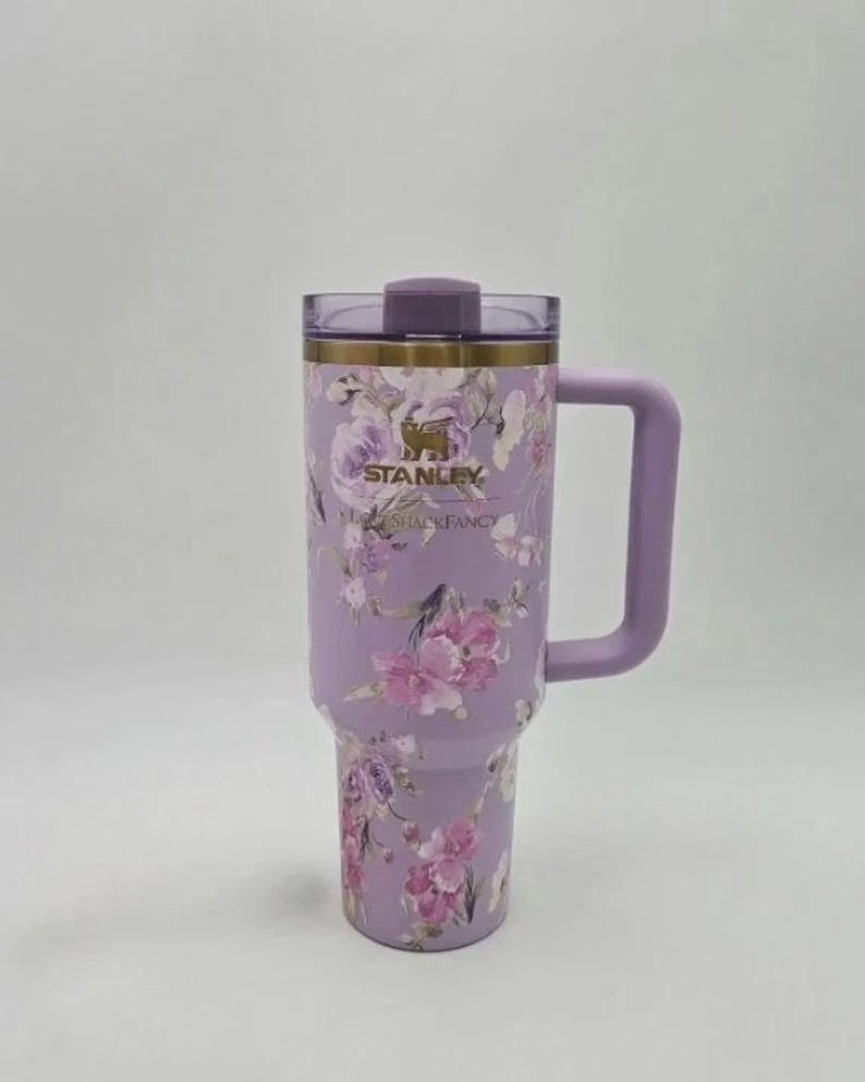 May include: A lavender stainless steel tumbler featuring a floral pattern with pink and purple roses. The cup has a gold-tone rim, a matching lavender handle, and a clear lid with a purple stopper. The Stanley and LoveShackFancy logos are printed in gold.