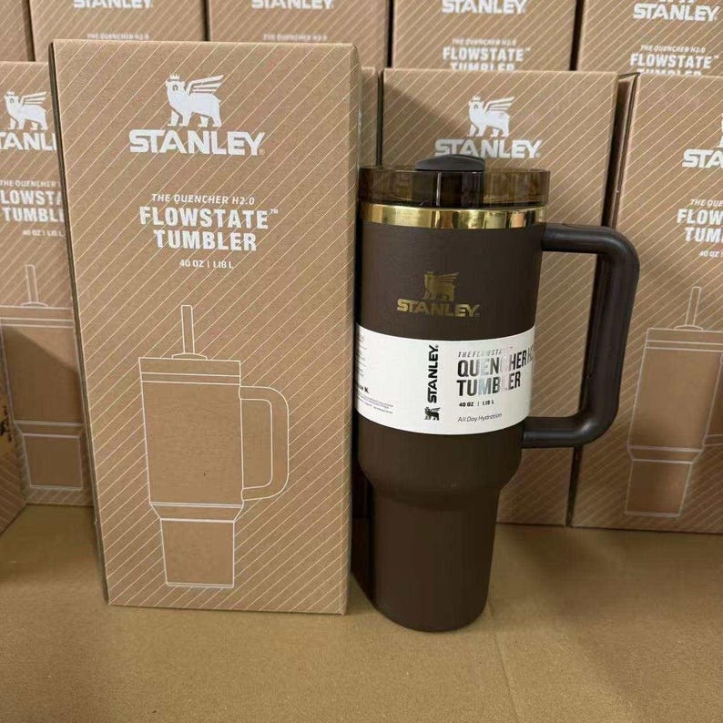 May include: A brown Stanley Flowstate tumbler with a handle and a straw, next to its cardboard box. The box features the Stanley logo and product details. The tumbler is 40 oz.