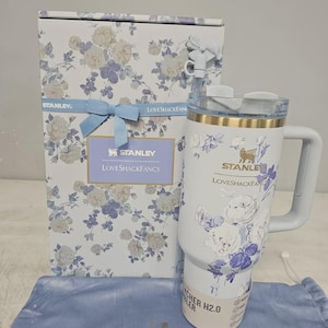 May include: A white Stanley Quencher H2.0 tumbler with a blue and white floral pattern, a gold rim, and a matching straw with a bow topper. It is displayed next to its rectangular box featuring the same floral print, a blue ribbon, and the text Stanley LoveShackFancy.