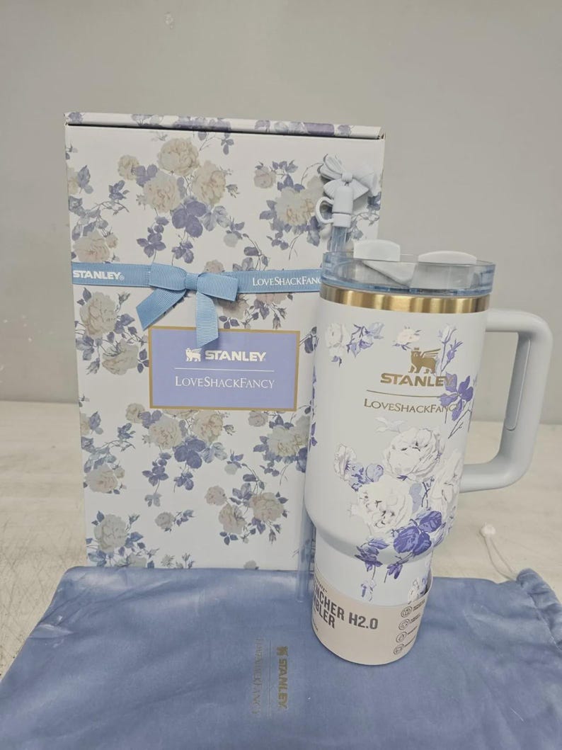 May include: A white Stanley Quencher H2.0 tumbler with a blue and white floral pattern, a gold rim, and a matching straw with a bow topper. It is displayed next to its rectangular box featuring the same floral print, a blue ribbon, and the text Stanley LoveShackFancy.