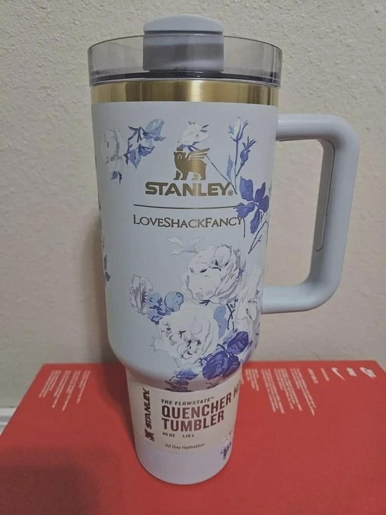 May include: A white Stanley Quencher tumbler with a blue floral pattern, a gold rim, and a clear lid. The front features the Stanley logo, the text LOVESHACKFANCY, and a label at the base reading THE FLOWSTATE QUENCHER TUMBLER 40 OZ 1.18 L.