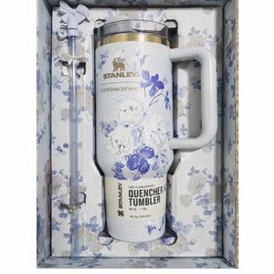 May include: A white 40 oz Stanley x LoveShackFancy Quencher tumbler with a blue and purple floral pattern, a side handle, a clear lid, and a matching straw with a bow topper, all packaged in a floral-patterned box with the product name printed.