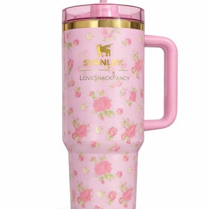 Pode incluir: Caneca Stanley rosa floral com al&ccedil;a. A caneca apresenta uma tampa e uma al&ccedil;a rosa, com detalhes dourados. O corpo da caneca &eacute; coberto por um padr&atilde;o floral rosa. O texto "STANLEY" e "LoveShackFancy" est&atilde;o impressos.