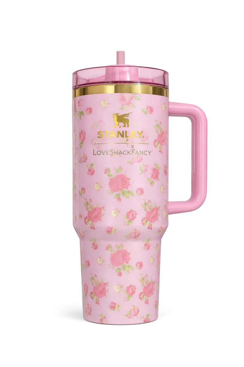 Pode incluir: Caneca Stanley rosa floral com al&ccedil;a. A caneca apresenta uma tampa e uma al&ccedil;a rosa, com detalhes dourados. O corpo da caneca &eacute; coberto por um padr&atilde;o floral rosa. O texto "STANLEY" e "LoveShackFancy" est&atilde;o impressos.