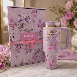 May include: A lavender Stanley tumbler with a floral pattern and a matching gift box. The tumbler features a gold Stanley logo, a handle, and a clear lid. The box is decorated with the same floral print, a pink ribbon, and a Stanley LoveShackFancy label.