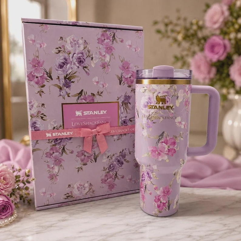 May include: A lavender Stanley tumbler with a floral pattern and a matching gift box. The tumbler features a gold Stanley logo, a handle, and a clear lid. The box is decorated with the same floral print, a pink ribbon, and a Stanley LoveShackFancy label.