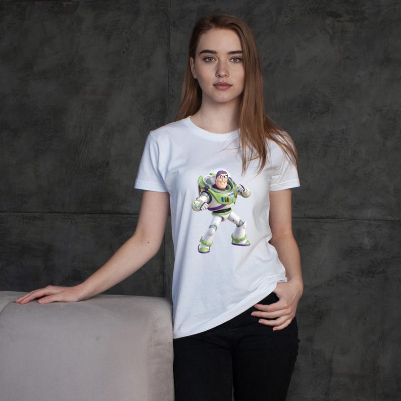 May include: White t-shirt featuring a graphic of Buzz Lightyear from Toy Story. The character is depicted in his signature space ranger uniform, with green, white, and purple accents. The shirt is a classic crew neck style.