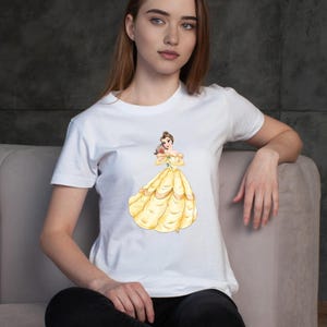 May include: White t-shirt featuring a watercolor illustration of a princess in a yellow gown holding a red rose. The princess has brown hair and is wearing a yellow dress with gold accents. The t-shirt is a classic crew neck style.