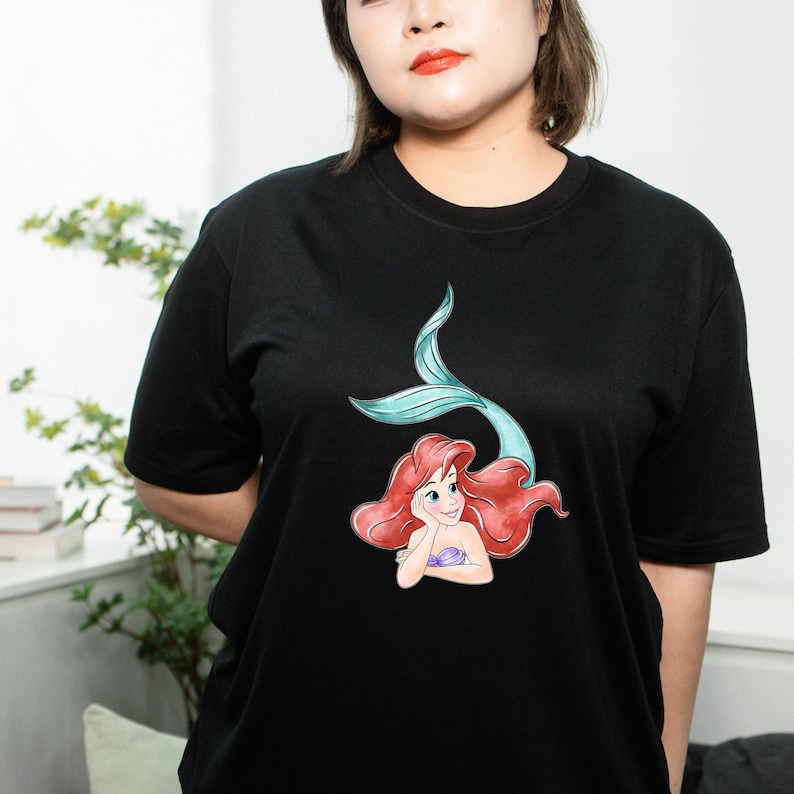 May include: Black t-shirt featuring a colorful graphic of a mermaid with red hair and a teal tail. The mermaid is resting her chin on her hand. The shirt is a classic crew neck style.
