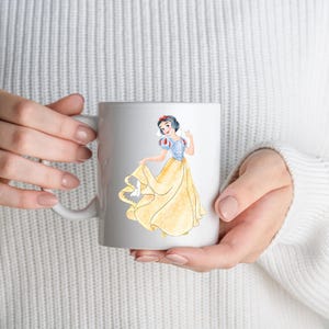 May include: White ceramic mug featuring a watercolor illustration of Snow White in her iconic yellow dress and blue bodice. The design includes a red bow in her hair. The mug is held by a person wearing a white sweater.