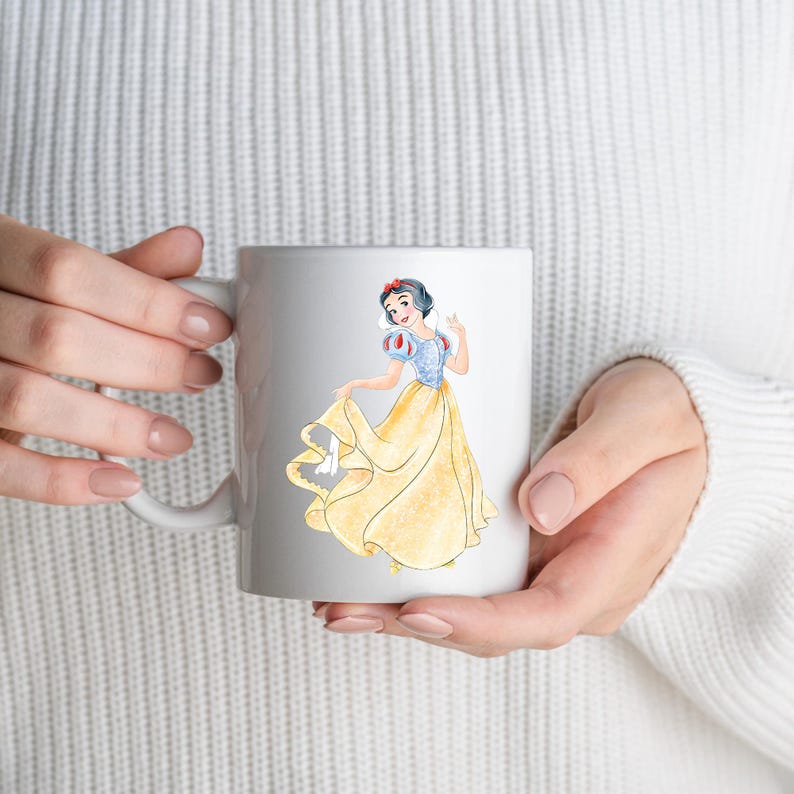 May include: White ceramic mug featuring a watercolor illustration of Snow White in her iconic yellow dress and blue bodice. The design includes a red bow in her hair. The mug is held by a person wearing a white sweater.
