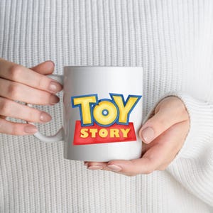 May include: White ceramic mug featuring the "Toy Story" logo in yellow and blue letters on a red background. The mug has a white handle and is held by a person wearing a white sweater.