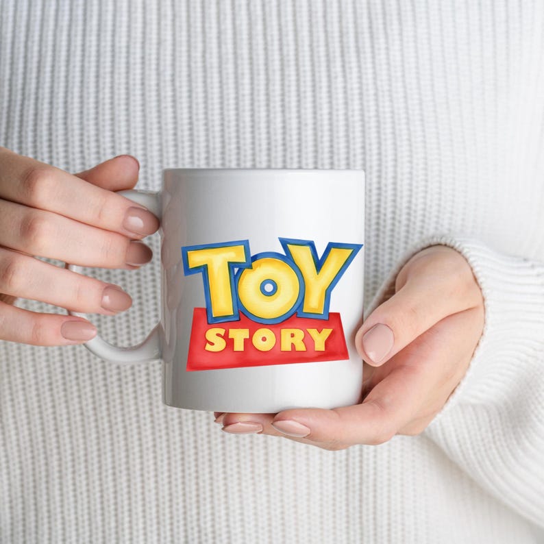 May include: White ceramic mug featuring the "Toy Story" logo in yellow and blue letters on a red background. The mug has a white handle and is held by a person wearing a white sweater.
