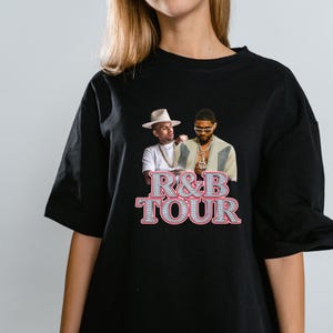 May include: Black t-shirt featuring a graphic of two men with the text "R&B TOUR" in a sparkling, gem-like font. The image shows the men in front of a light background. The t-shirt is a classic crew neck style.