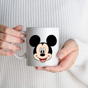 Op de afbeelding: Witte keramische mok met een lachend Mickey Mouse-ontwerp. Het stripfiguur heeft grote zwarte oren, een zwarte neus en een rode mond. De mok wordt vastgehouden door een persoon die een witte trui draagt.