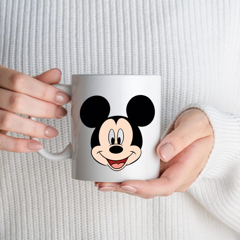 Op de afbeelding: Witte keramische mok met een lachend Mickey Mouse-ontwerp. Het stripfiguur heeft grote zwarte oren, een zwarte neus en een rode mond. De mok wordt vastgehouden door een persoon die een witte trui draagt.