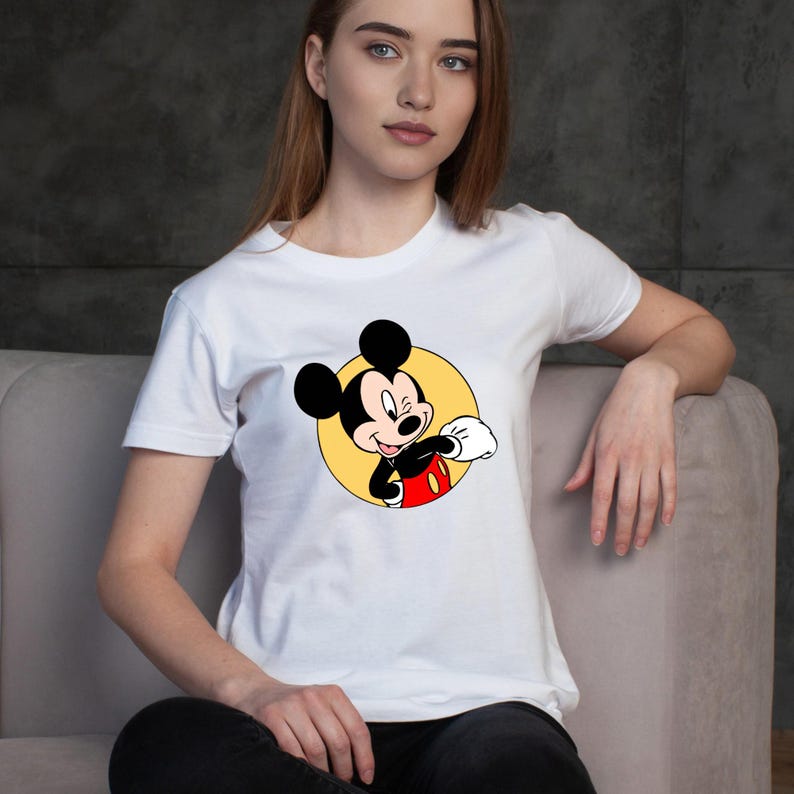 Op de afbeelding: Wit T-shirt met een cartoonafbeelding van Mickey Mouse. De afbeelding toont Mickey Mouse die knipoogt, in een gele cirkel. Hij draagt een zwart jasje, een rode broek en witte handschoenen. Het T-shirt heeft een ronde hals.