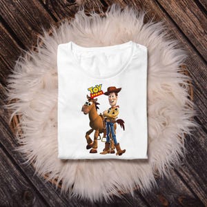 May include: White t-shirt featuring a graphic of Woody from Toy Story with his horse. The Toy Story logo is above the characters. The shirt is folded and placed on a fluffy, white surface.