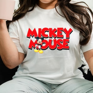 Op de afbeelding: Wit T-shirt met de tekst "MICKEY MOUSE" in het rood met een zwarte omlijning. Het iconische Mickey Mouse-personage staat onder de tekst afgebeeld, met een rode broek, gele schoenen en witte handschoenen.