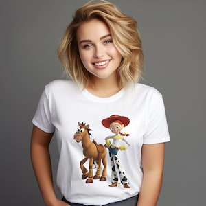 May include: White t-shirt featuring a cartoon illustration of a horse and a cowgirl character. The cowgirl wears a red hat, yellow shirt, blue jeans, and black and white patterned pants. The horse has a brown saddle.