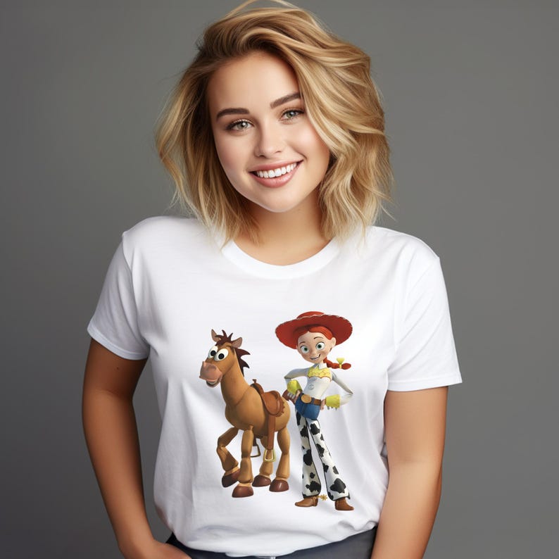 May include: White t-shirt featuring a cartoon illustration of a horse and a cowgirl character. The cowgirl wears a red hat, yellow shirt, blue jeans, and black and white patterned pants. The horse has a brown saddle.
