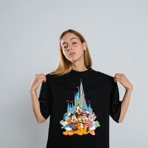 Op de afbeelding: Zwart t-shirt met een kleurrijke afbeelding van Disney-figuren voor een kasteel. De afbeelding bevat Mickey Mouse, Minnie Mouse, Donald Duck, Goofy en Daisy Duck. Het kasteel heeft blauwe torens en rode vlaggen.