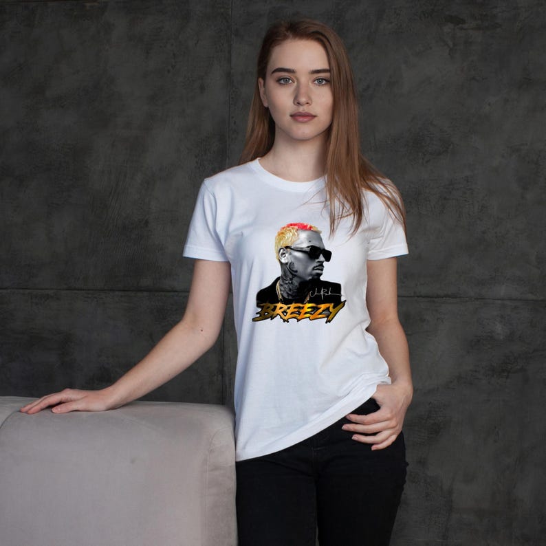May include: White t-shirt featuring a graphic of a person with sunglasses and the word "BREEZY" in gold and black. The person in the graphic has blonde and red hair. The shirt is being worn by a person.