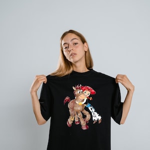 May include: Black t-shirt featuring a cartoon illustration of a girl with red hair hugging a brown horse. The girl is wearing a blue and white outfit. The horse has a yellow collar. The t-shirt is a solid black color.
